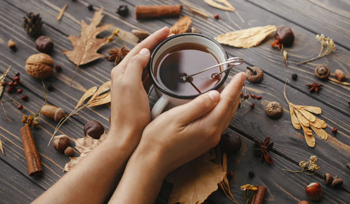 Manos en reposo sobre una mesa de madera clara, junto a una taza de té y un cuaderno abierto, con luz natural suave