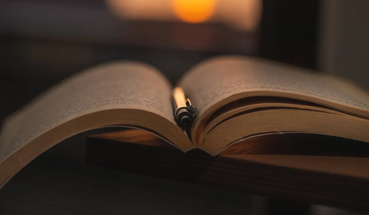 Libro abierto con una pluma estilográfica entre las páginas, sobre mesa de madera oscura, con luz cálida de una vela al fondo