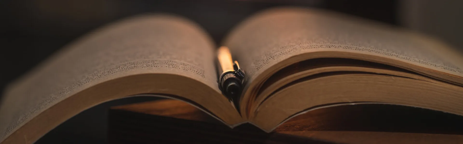 Libro abierto con una pluma estilográfica entre las páginas, sobre mesa de madera oscura, con luz cálida de una vela al fondo