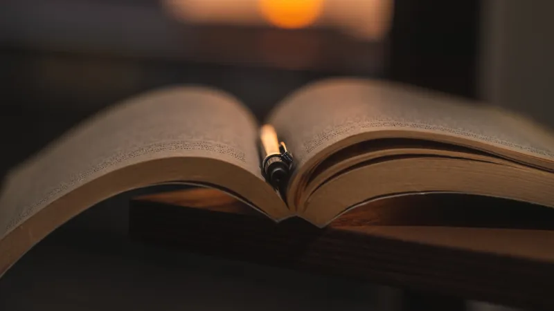 Libro abierto con una pluma estilográfica entre las páginas, sobre mesa de madera oscura, con luz cálida de una vela al fondo